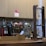 A coffee shop counter with a large espresso machine and a siphon coffee maker. The espresso machine is stainless steel with red accents and several knobs and dials. A wooden structure on the right holds glass vessels for brewing coffee. The background features light-colored cabinetry and a backsplash of small, round tiles in neutral tones.