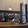 A coffee shop counter with a large espresso machine and a siphon coffee maker. The espresso machine is stainless steel with red accents and several knobs and dials. A wooden structure on the right holds glass vessels for brewing coffee. The background features light-colored cabinetry and a backsplash of small, round tiles in neutral tones.