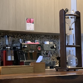 A coffee shop counter with a large espresso machine and a siphon coffee maker. The espresso machine is stainless steel with red accents and several knobs and dials. A wooden structure on the right holds glass vessels for brewing coffee. The background features light-colored cabinetry and a backsplash of small, round tiles in neutral tones.