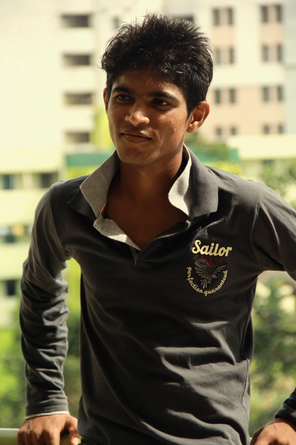 In the image, there is a young man standing against a backdrop of a cityscape. He is wearing a gray long-sleeved shirt with a white collar and a black tie around his neck. The shirt features a logo on the left chest area that reads "Sailor". His hair is styled in a short cut, giving him a casual yet stylish appearance.

The man's gaze is directed off to the side, suggesting he might be observing something or someone outside of the frame.
