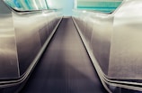 A futuristic perspective of a moving walkway with shiny stainless steel sides, leading towards a tiled wall with a blue and teal gradient. The walkway surface is black with parallel ridges running its length.