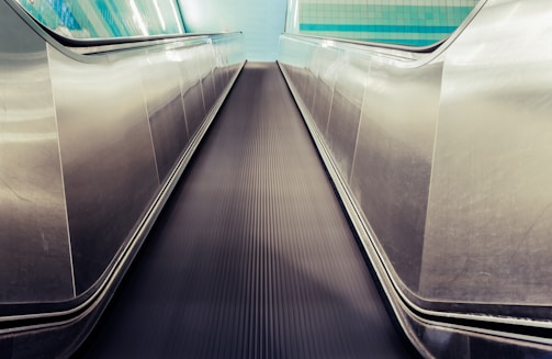 A futuristic perspective of a moving walkway with shiny stainless steel sides, leading towards a tiled wall with a blue and teal gradient. The walkway surface is black with parallel ridges running its length.