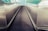 A futuristic perspective of a moving walkway with shiny stainless steel sides, leading towards a tiled wall with a blue and teal gradient. The walkway surface is black with parallel ridges running its length.