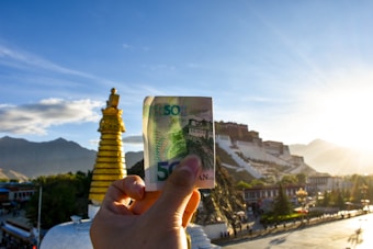 A hand holds a banknote in the foreground, with a golden religious stupa and a grand white palace on a hill in the background. The setting sun casts a warm glow over the scene, with mountains visible in the distance.