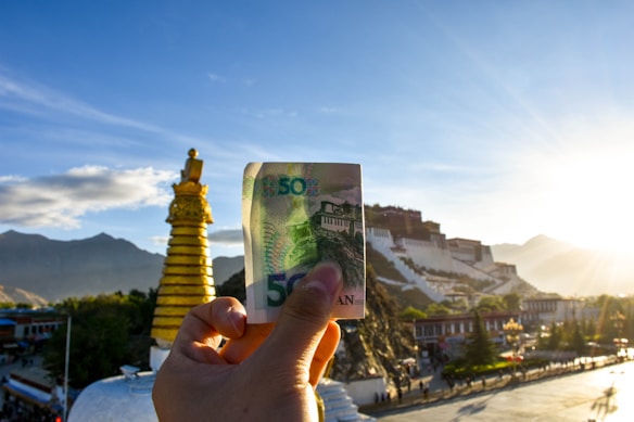 A hand holds a banknote in the foreground, with a golden religious stupa and a grand white palace on a hill in the background. The setting sun casts a warm glow over the scene, with mountains visible in the distance.