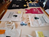 On a wooden table, various items are neatly displayed. Two tote bags with the phrase 'Feedback is the Breakfast of Champions' are featured prominently. Several casual clothing items, including a black T-shirt and a pink tank top, are laid out. Each item has a small note attached, seemingly indicating names or labels. A toy truck with a blue and white pattern sits among the items.