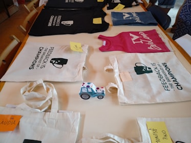 On a wooden table, various items are neatly displayed. Two tote bags with the phrase 'Feedback is the Breakfast of Champions' are featured prominently. Several casual clothing items, including a black T-shirt and a pink tank top, are laid out. Each item has a small note attached, seemingly indicating names or labels. A toy truck with a blue and white pattern sits among the items.
