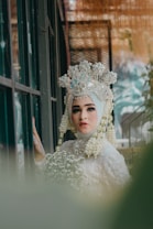 A woman in traditional bridal attire stands with a serene expression, wearing an ornate, floral headdress adorned with jewels and white flowers. Her attire is richly decorated, and she holds a small bouquet of white flowers. The background has a soft, blurred effect with greenery, adding to the dreamlike quality.