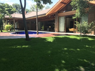 A modern house with a sloped roof and large windows sits amidst a lush garden. A wooden deck surrounds a small pool, and the area is shaded by tall trees.