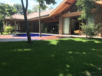 A modern house with a sloped roof and large windows sits amidst a lush garden. A wooden deck surrounds a small pool, and the area is shaded by tall trees.