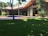 A modern house with a sloped roof and large windows sits amidst a lush garden. A wooden deck surrounds a small pool, and the area is shaded by tall trees.