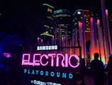 An outdoor night scene features a vibrant and illuminated sign reading 'Samsung Electric Playground' in neon colors. The background includes modern buildings and numerous tall, colorful light installations. A few people are visible, and the atmosphere is lively with bright, colorful lights dominating the setting.