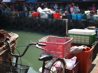 A bicycle with a red basket is positioned near a dockside with various colorful plastic crates and containers stacked in the background. Several people are faintly visible and appear to be involved in market activities or trades. The scene has an industrial and busy marketplace atmosphere, with tires lining the dock.