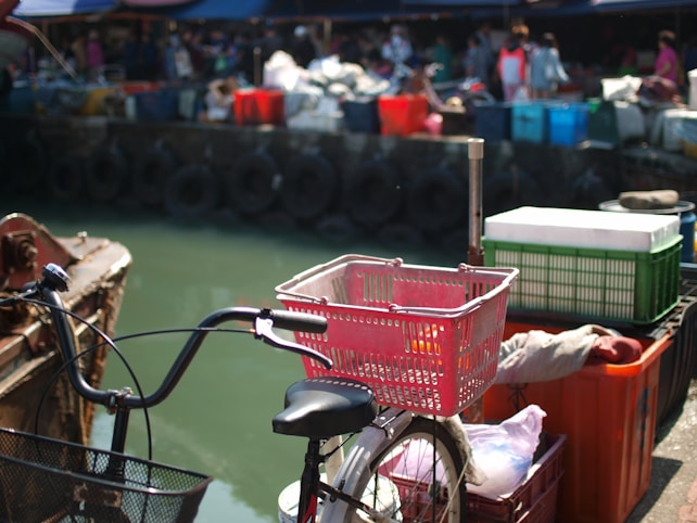 A bicycle with a red basket is positioned near a dockside with various colorful plastic crates and containers stacked in the background. Several people are faintly visible and appear to be involved in market activities or trades. The scene has an industrial and busy marketplace atmosphere, with tires lining the dock.