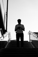 A silhouette of a person standing at the top of a staircase, holding a camera. The image is in black and white, with the person in a striped shirt and a cap. Glass railings and architecture are visible on the sides, suggesting an outdoor urban setting.