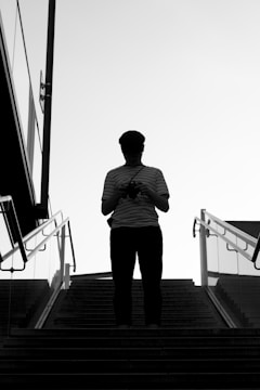 A silhouette of a person standing at the top of a staircase, holding a camera. The image is in black and white, with the person in a striped shirt and a cap. Glass railings and architecture are visible on the sides, suggesting an outdoor urban setting.