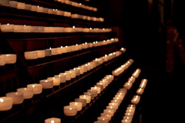 Rows of small, white candles are arranged in a dark, dimly lit space, creating a serene and tranquil atmosphere. The candles emit a soft, warm glow, illuminating their surroundings.