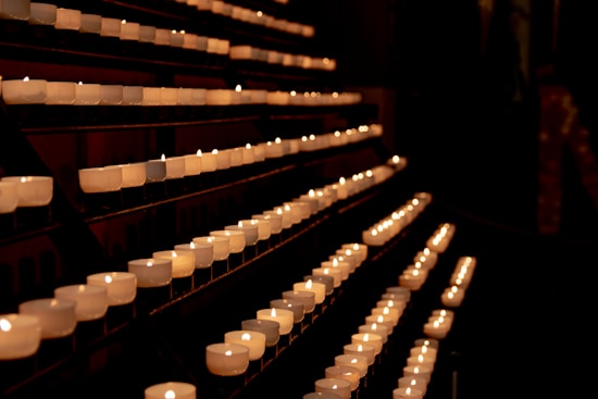 Rows of small, white candles are arranged in a dark, dimly lit space, creating a serene and tranquil atmosphere. The candles emit a soft, warm glow, illuminating their surroundings.