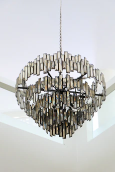 A large, ornate chandelier made of numerous rectangular, metallic pieces arranged in a circular, tiered pattern, suspended from the ceiling by a long chain. The metal reflects light, creating a subtle interplay of shadows and highlights. The background is a minimalistic white, emphasizing the chandelier's intricate design.