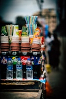 A collection of bottled drinks including popular soda brands like Coca-Cola, Sprite, and Thums Up is displayed. The bottles are accompanied by stacks of paper cups placed on top, with colorful straws protruding from one of the cups. The setup appears to be outdoors, possibly in a street setting.