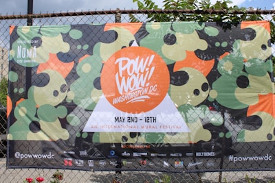 A colorful festival banner hung on a chain-link fence, promoting the Pow! Wow! Washington DC event. The design features abstract shapes in shades of green, black, and orange. The text includes event dates and location, along with sponsor logos at the bottom.