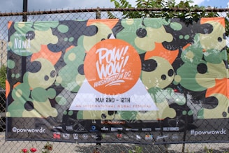 A colorful festival banner hung on a chain-link fence, promoting the Pow! Wow! Washington DC event. The design features abstract shapes in shades of green, black, and orange. The text includes event dates and location, along with sponsor logos at the bottom.