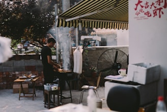 A person is grilling food on a barbecue in an outdoor area with a striped awning overhead. There's a fan nearby, a small table with items on it, and a wall decorated with colorful bricks in the background. The area appears casual and relaxed, with some smoke rising from the grill.