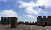 A stone fortress with crenellated walls stands under a bright blue sky. A Union Jack flag flutters on a flagpole, adding a vibrant contrast to the historic stonework. The courtyard is paved with bricks and features a tall brick structure and a small entrance in one of the castle walls.