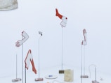 Abstract sculptures resembling disassembled body parts, such as arms, hands, and legs, are mounted on slender stands against a minimalistic, light-colored background. The artwork consists of distinct geometric shapes in red and white, evoking a sense of surrealism. Blocks and an abstract curling form rest on the floor among the sculptures.