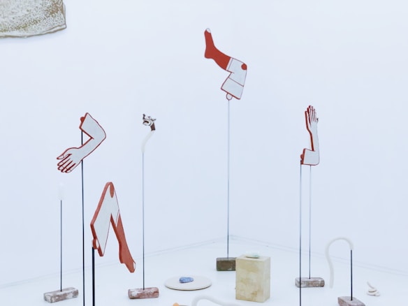 Abstract sculptures resembling disassembled body parts, such as arms, hands, and legs, are mounted on slender stands against a minimalistic, light-colored background. The artwork consists of distinct geometric shapes in red and white, evoking a sense of surrealism. Blocks and an abstract curling form rest on the floor among the sculptures.