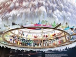 A circular display containing numerous bottles of liquor arranged on shelves. The shelves are encircled by decorative white feathers creating a unique and artistic presentation. A range of bottles featuring diverse shapes and colors is presented, indicating a wide selection of spirits. Below the display, there is a textual description about 'Angel's Share', explaining its significance in the context of distilled beverages.