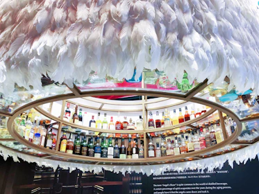 A circular display containing numerous bottles of liquor arranged on shelves. The shelves are encircled by decorative white feathers creating a unique and artistic presentation. A range of bottles featuring diverse shapes and colors is presented, indicating a wide selection of spirits. Below the display, there is a textual description about 'Angel's Share', explaining its significance in the context of distilled beverages.
