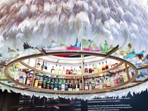 A circular display containing numerous bottles of liquor arranged on shelves. The shelves are encircled by decorative white feathers creating a unique and artistic presentation. A range of bottles featuring diverse shapes and colors is presented, indicating a wide selection of spirits. Below the display, there is a textual description about 'Angel's Share', explaining its significance in the context of distilled beverages.