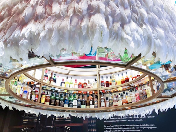 A circular display containing numerous bottles of liquor arranged on shelves. The shelves are encircled by decorative white feathers creating a unique and artistic presentation. A range of bottles featuring diverse shapes and colors is presented, indicating a wide selection of spirits. Below the display, there is a textual description about 'Angel's Share', explaining its significance in the context of distilled beverages.