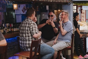 Three men are sitting at a bar, engaging in a lively conversation. One man is holding a glass of beer, while another is contemplating with his hand on his chin. The bar has a variety of bottled drinks on the counter and is decorated with various posters and signs. In the background, a woman is preparing food at a street food stall.