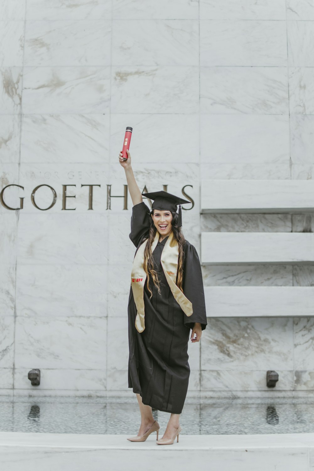 mulher sorridente em pé e vestindo argamassa e vestido acadêmico levantando o braço direito com diploma vermelho
