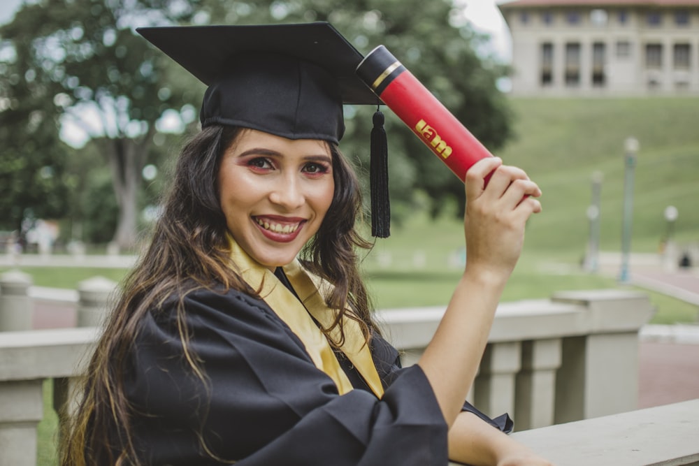 smiling woman wearing mortar board and academic gown holding red and black diploma