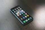 A smartphone with a variety of apps displayed on its screen, placed on a dark wooden surface. The screen shows a mix of social media, entertainment, and utility apps, neatly organized into folders and individual icons.