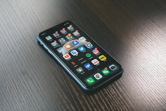 A smartphone with a variety of apps displayed on its screen, placed on a dark wooden surface. The screen shows a mix of social media, entertainment, and utility apps, neatly organized into folders and individual icons.
