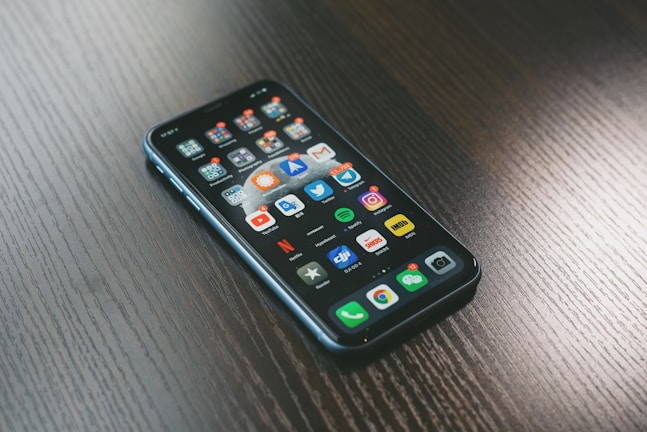 A smartphone with a variety of apps displayed on its screen, placed on a dark wooden surface. The screen shows a mix of social media, entertainment, and utility apps, neatly organized into folders and individual icons.