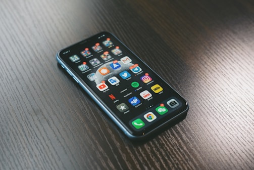 A smartphone with a variety of apps displayed on its screen, placed on a dark wooden surface. The screen shows a mix of social media, entertainment, and utility apps, neatly organized into folders and individual icons.