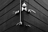 A selection of security cameras are mounted on a brick wall, arranged at the corner of the building. The wall is composed of dark bricks, creating a stark contrast with the white and metallic colors of the cameras. The layout includes various types of cameras, each pointing in different directions.