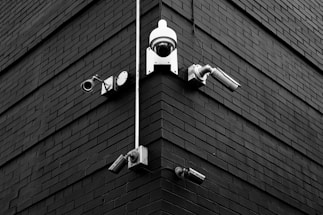 A selection of security cameras are mounted on a brick wall, arranged at the corner of the building. The wall is composed of dark bricks, creating a stark contrast with the white and metallic colors of the cameras. The layout includes various types of cameras, each pointing in different directions.