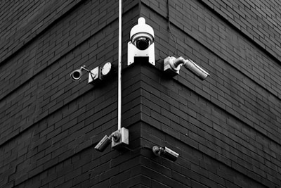 A selection of security cameras are mounted on a brick wall, arranged at the corner of the building. The wall is composed of dark bricks, creating a stark contrast with the white and metallic colors of the cameras. The layout includes various types of cameras, each pointing in different directions.