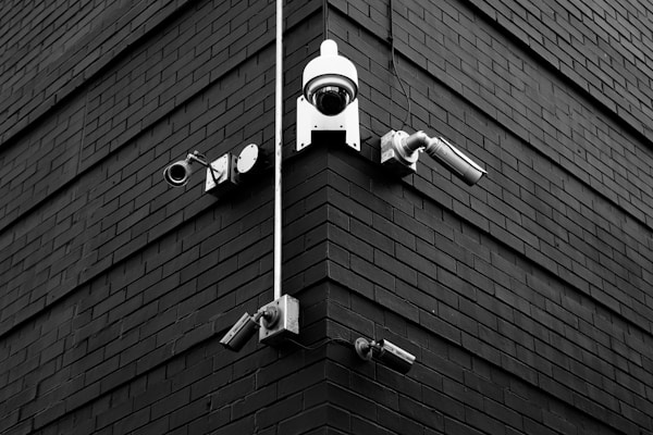 A selection of security cameras are mounted on a brick wall, arranged at the corner of the building. The wall is composed of dark bricks, creating a stark contrast with the white and metallic colors of the cameras. The layout includes various types of cameras, each pointing in different directions.