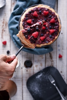 A hand holding a slice of berry-topped cheesecake with visible raspberries and blueberries. The cheesecake sits on a rustic wooden table, garnished with more berries. A fork and a scattering of powdered sugar are seen nearby.