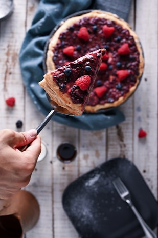 A hand holding a slice of berry-topped cheesecake with visible raspberries and blueberries. The cheesecake sits on a rustic wooden table, garnished with more berries. A fork and a scattering of powdered sugar are seen nearby.