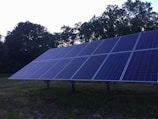 A large array of solar panels is installed outdoors with a backdrop of trees. The sky appears to be overcast, and the panels are set on a grassy environment. The structure supporting the panels is clearly visible.