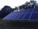 A large array of solar panels is installed outdoors with a backdrop of trees. The sky appears to be overcast, and the panels are set on a grassy environment. The structure supporting the panels is clearly visible.