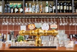 A bar setting featuring a polished gold beer tap with various labeled handles. Behind the bar, there are shelves stocked with bottles of wine and spirits, along with neatly hanging wine glasses. The countertop displays several clean glasses and there are elements of green plants and fruit adding color to the scene.